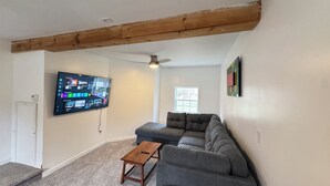 Un soggiorno con un divano componibile, un tavolino in legno e una TV a parete che mostra l'interfaccia di un servizio di streaming.