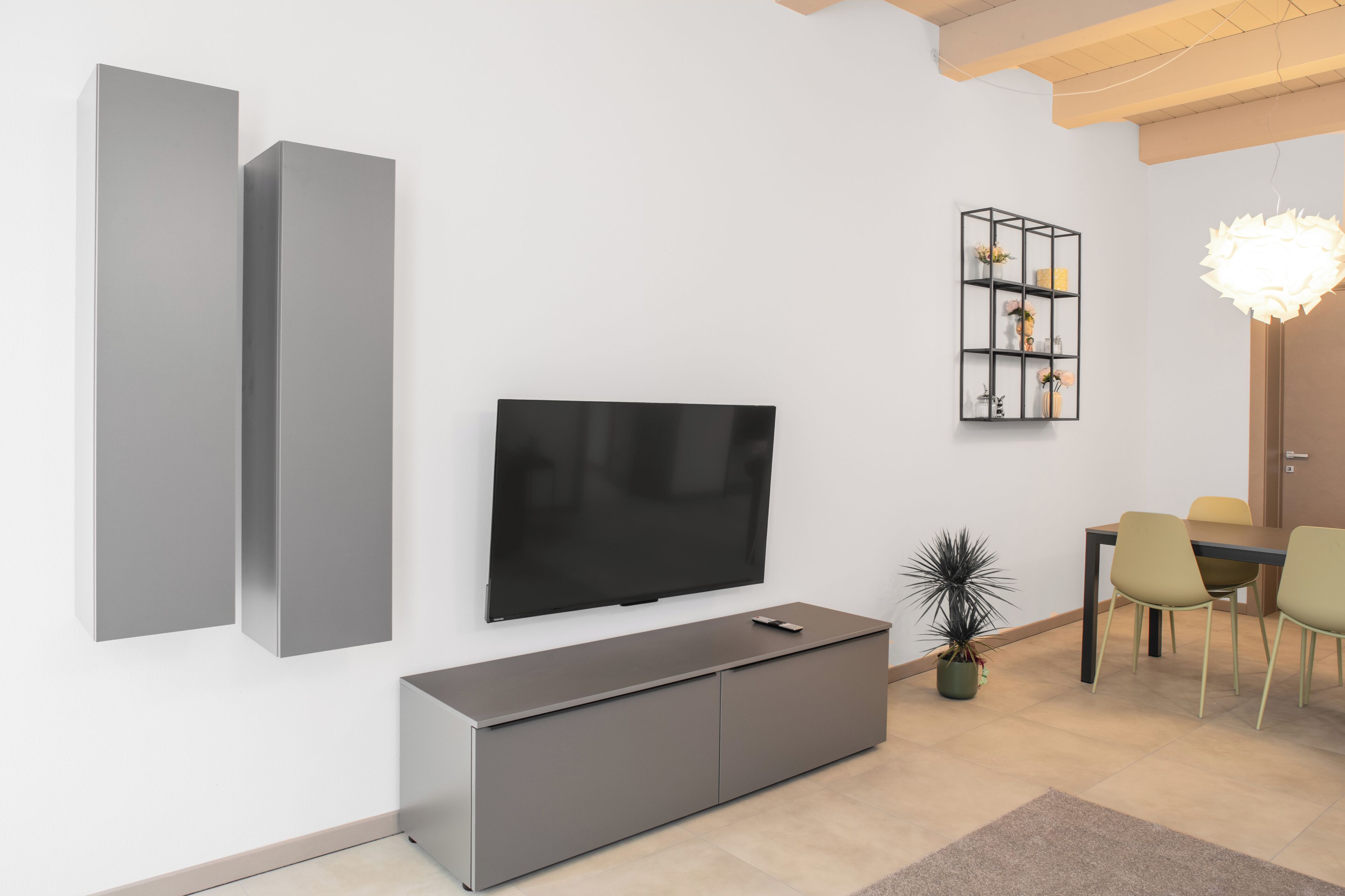 A modern living room with a flat-screen TV, grey cabinets, a dining area with chairs, and a decorative plant.