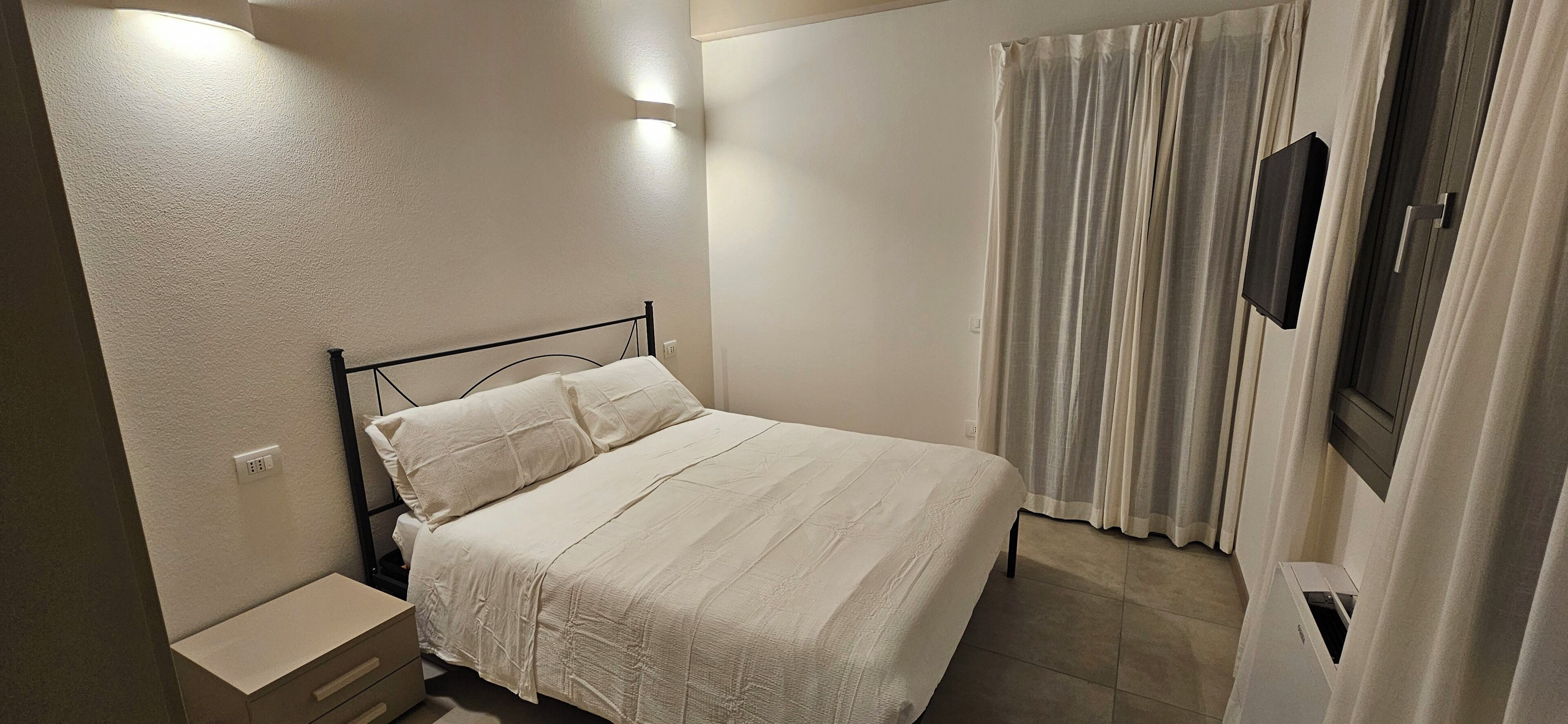 A neatly made bed with a white bedspread and pillows, a bedside table with a lamp, and a wall-mounted TV.
