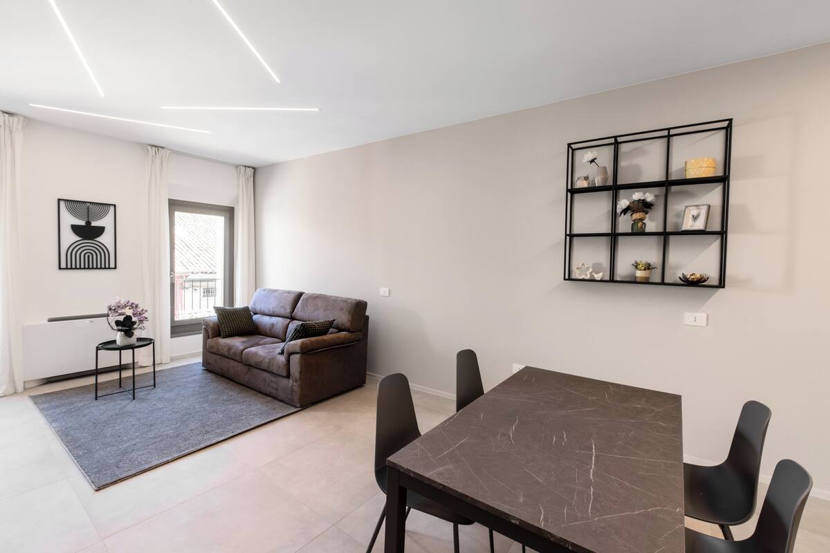 A modern living room with a brown leather sofa, a dining table with black chairs, a grey rug, and a black shelving unit on the wall.