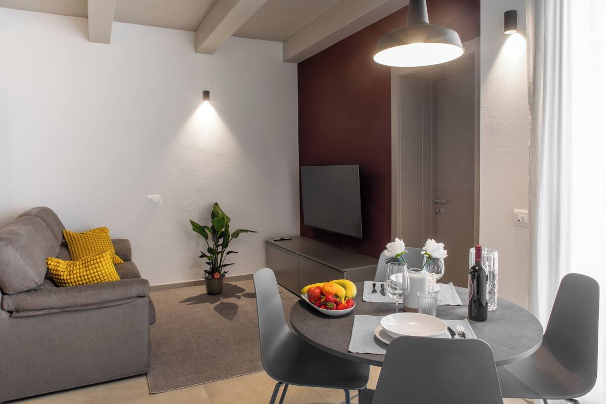 A modern living room with a gray sofa, a dining table with fruit, wine, and glasses, and a flat-screen TV mounted on the wall.