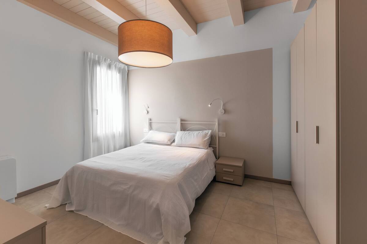A bedroom with a bed, bedside tables, a nightstand, a hanging light fixture, and a window with curtains.