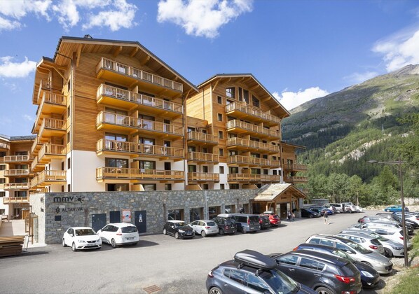 Un edificio de madera de varios pisos con balcones, un estacionamiento y montañas al fondo.