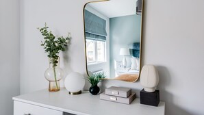 A white dresser with a mirror, a vase with green plants, a round white lamp, and a small black vase.