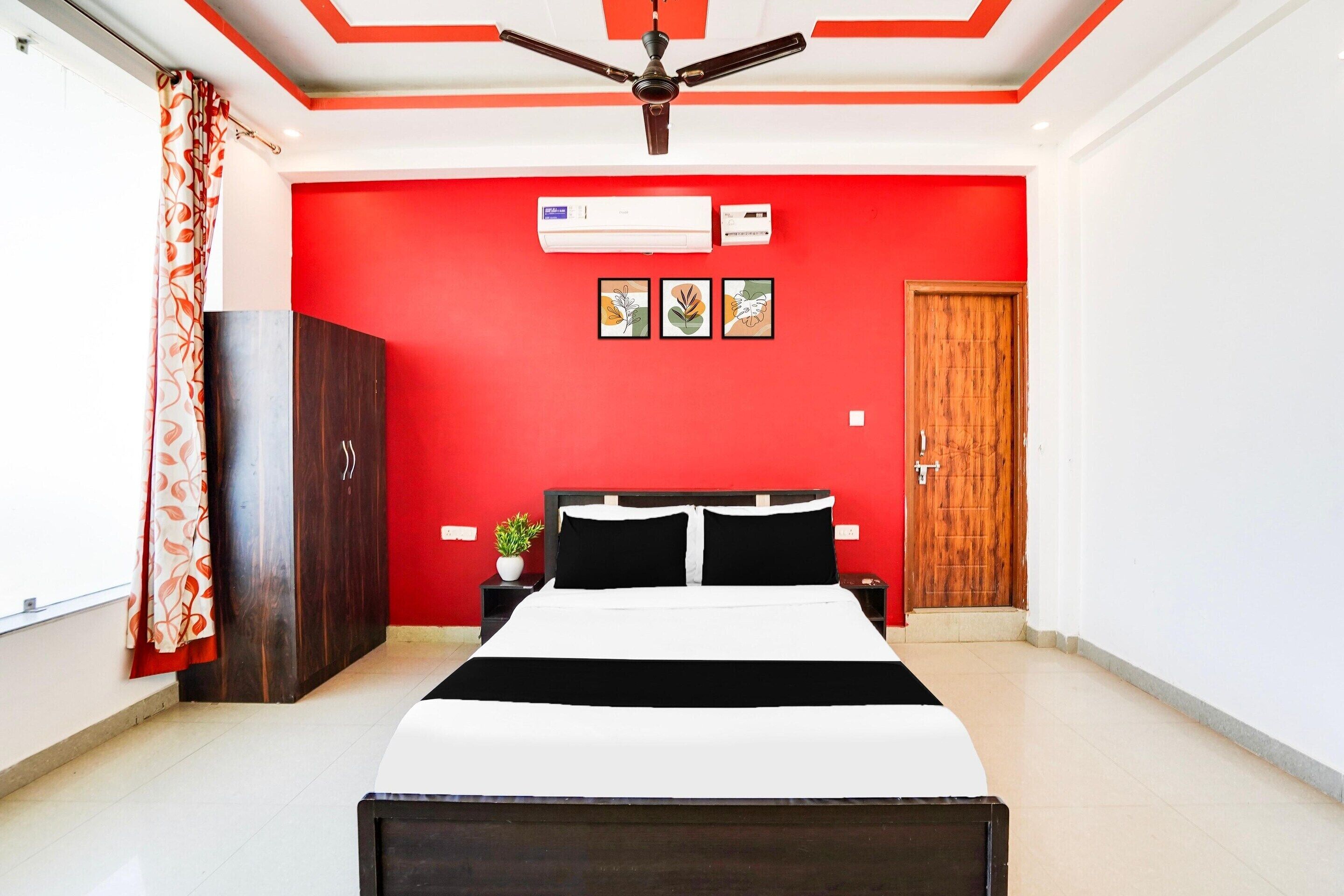A bedroom with a bed, a ceiling fan, an air conditioning unit, a wooden wardrobe, a nightstand, a potted plant, and a door.