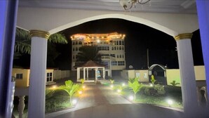 A hotel entrance with a covered walkway, palm trees, and a well-lit building in the background. - Chateau Mbeka'a Paris (Kribi)