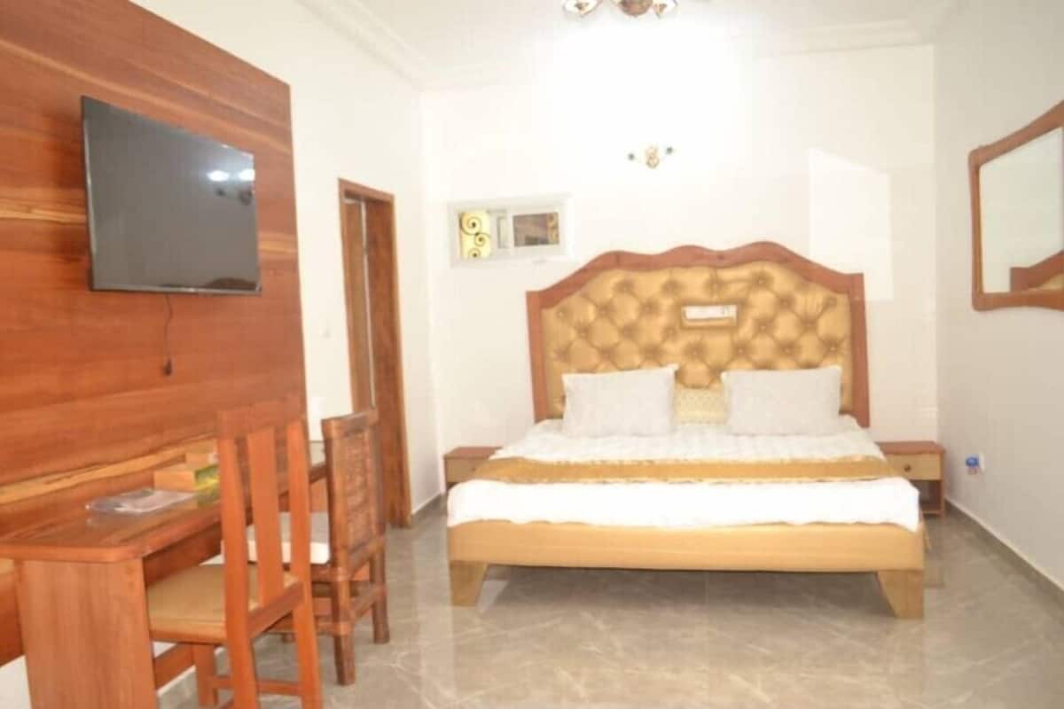A bedroom with a wooden headboard, a bed with white bedding, a television mounted on the wall, and a small window with decorative curtains.