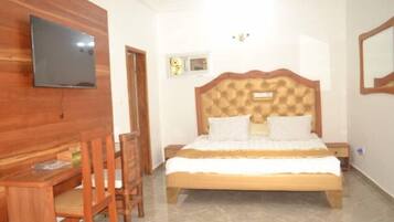 Un dormitorio con cabecera de madera, cama con ropa blanca, televisor montado en la pared y un ventanal con cortinas decorativas.
