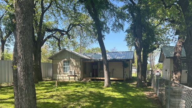 Une petite maison de plain-pied avec un porche, entourée d’une clôture en treillis métallique et d’arbres.