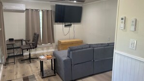 Un salon avec un canapé gris, une petite table, une chaise, un téléviseur fixé au mur et un ventilateur de plafond.