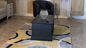 Una sala de estar con ventilador de techo, un sillón negro, un jarrón de vidrio, una lámpara y una alfombra estampada.