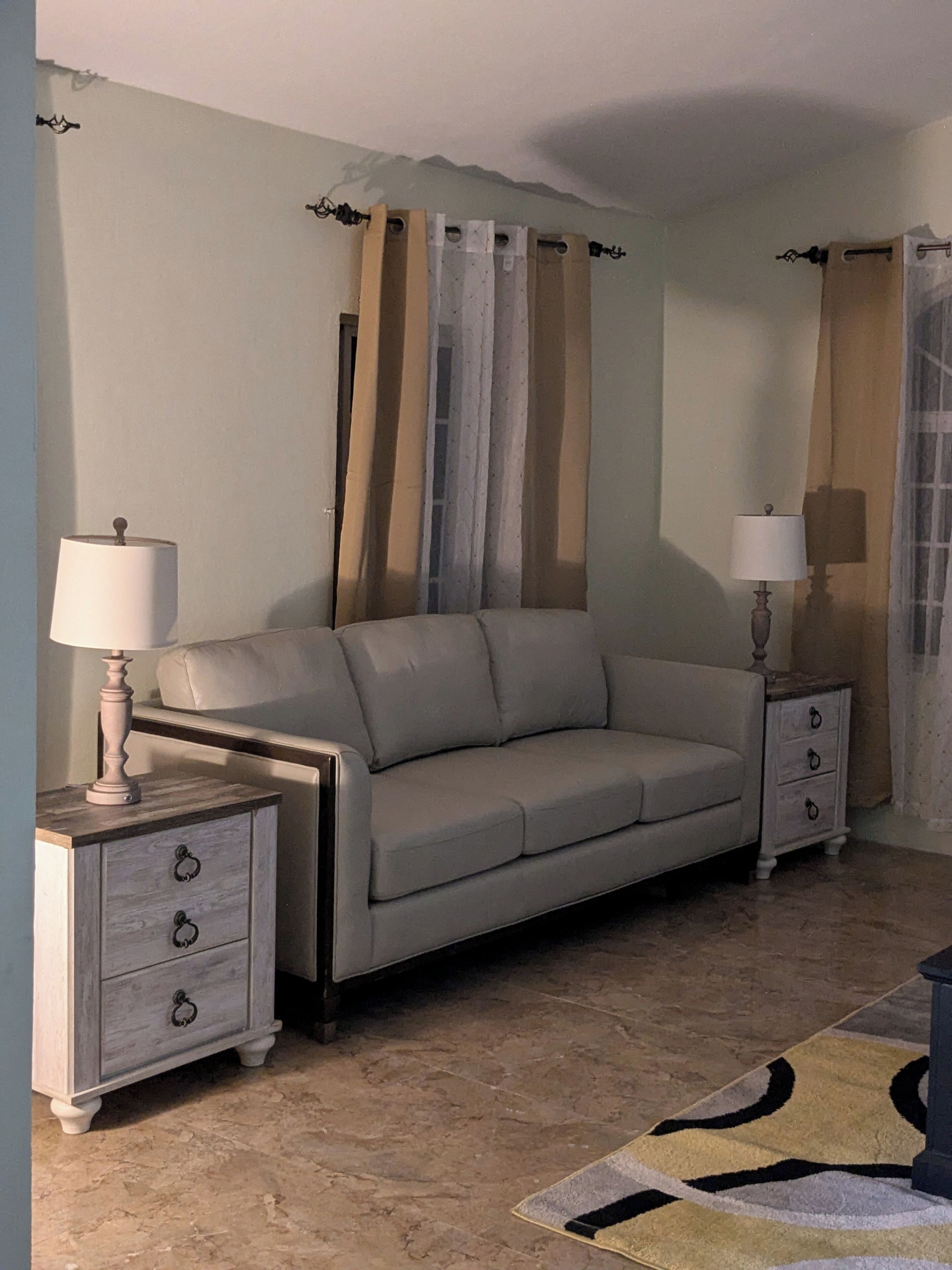 Una sala de estar con un sofá, dos mesitas de noche con lámparas, un tapete estampado y cortinas.