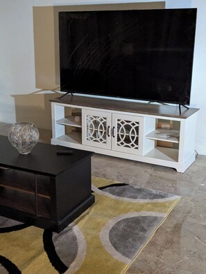 Una sala de estar con un televisor de pantalla plana sobre un mueble blanco, un aparador de madera oscura y una alfombra estampada.