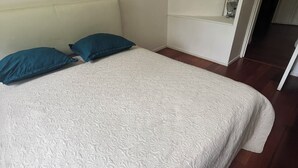 Un lit bien fait, avec une couvre-lit à motifs et deux oreillers, une table de chevet avec une lampe, et une porte donnant sur une autre pièce.