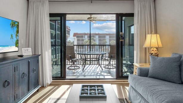 Una sala moderna con sofá, televisión, mesa de comedor con sillas y un balcón con vistas a edificios y palmeras.