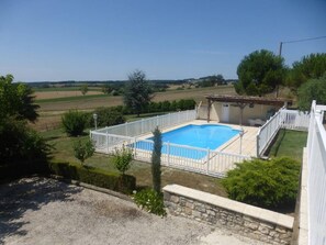 Une piscine rectangulaire entourée d’une clôture blanche, elle-même bordée d’un mur de pierre et d’une zone herbeuse.