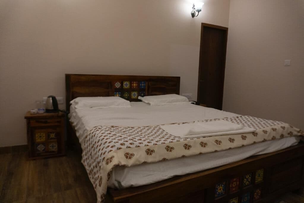 A neatly made bed with a patterned bedspread and pillows, a wooden headboard, a nightstand with a decorative box, and a wall-mounted light fixture.