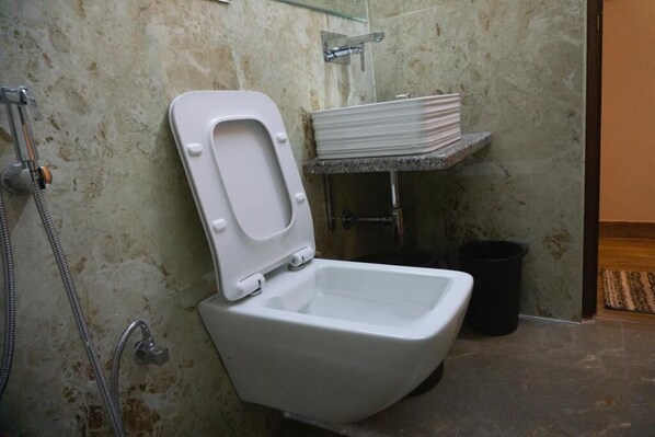 Un inodoro blanco con asiento elevado, un cabezal de ducha, una pila de platos blancos y un cubo de basura negro en un baño con pared texturizada.