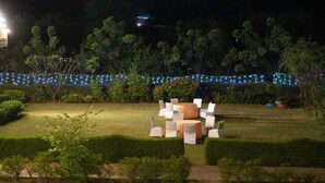 A well-lit outdoor seating area with round tables and chairs, surrounded by neatly trimmed hedges and trees, under a string of blue lights. - Sultanpur Resorts (Gurugram)