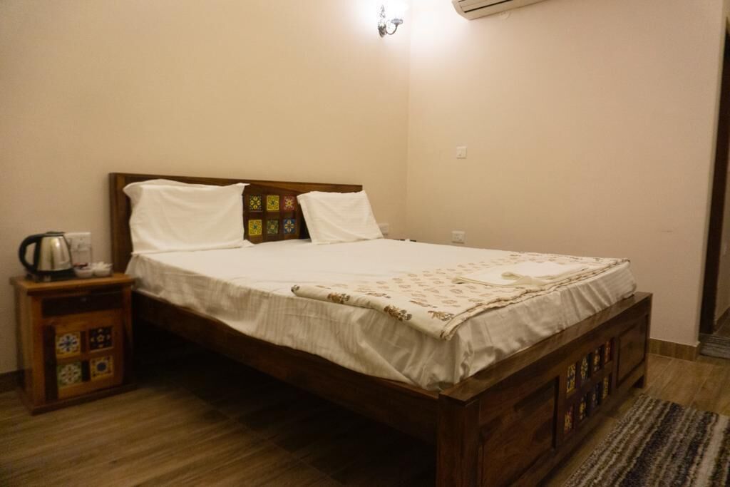 A single bed with a wooden frame and a white bedspread, a nightstand with a kettle, and a wall-mounted air conditioner.