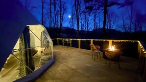 A geodesic dome structure with a fire pit and seating area on a wooden deck, surrounded by trees and string lights.
