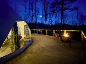 Eine geodätische Kuppel mit Feuerstelle und Sitzgelegenheit auf einer Holzterrasse, umgeben von Bäumen und Lichterketten.