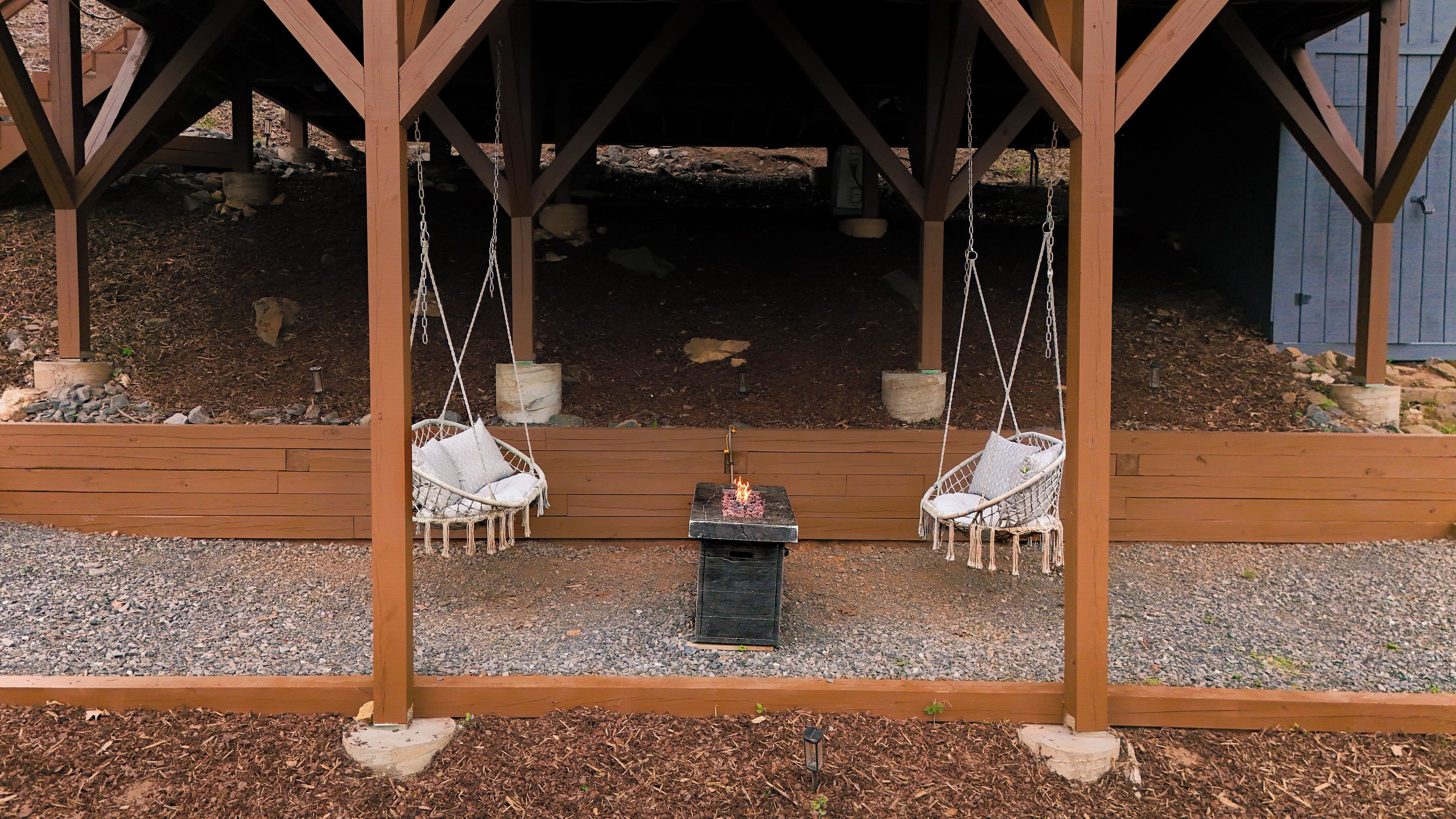 A wooden swing set with two swings, a small fire pit in the center, and a gravel ground.