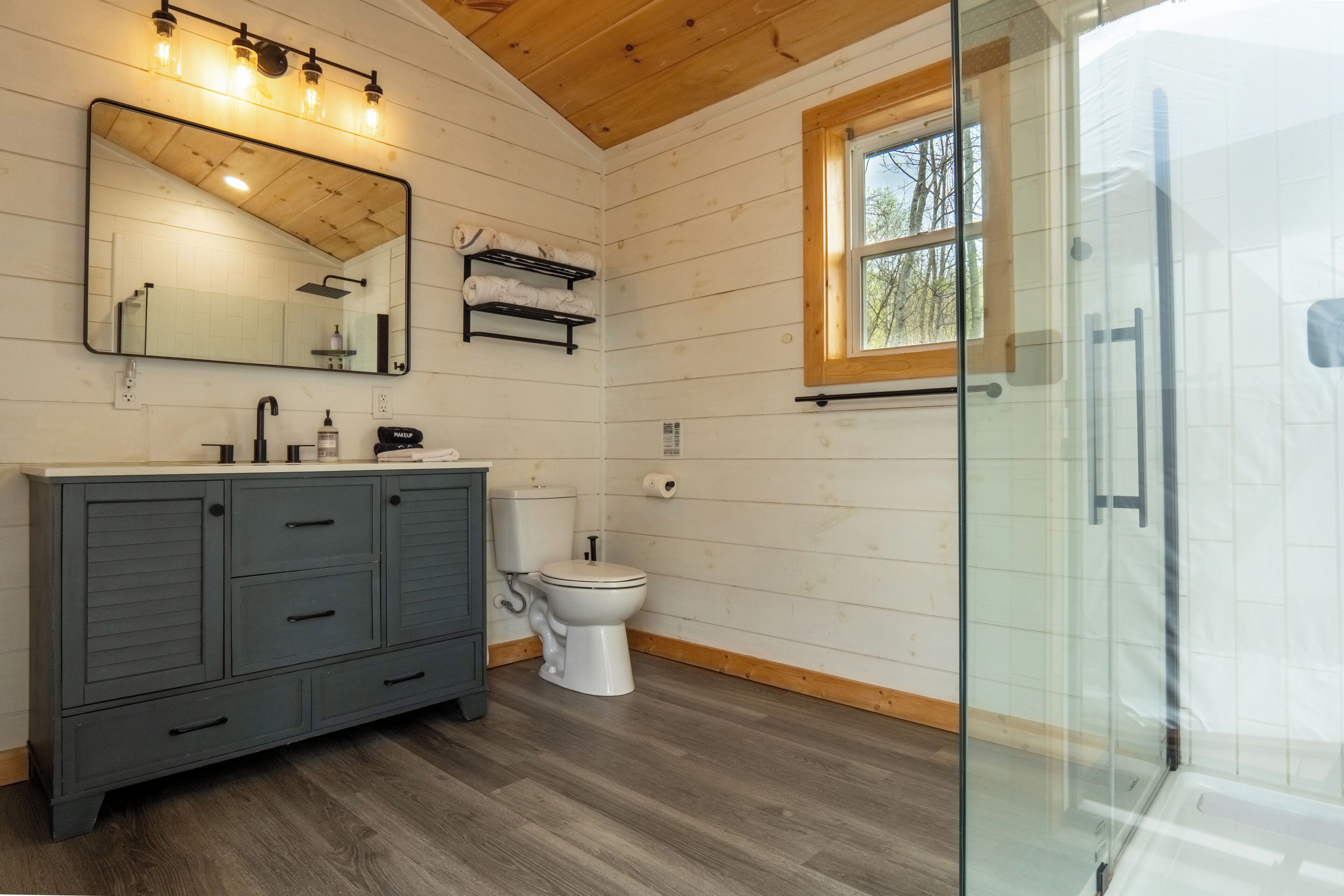 Une salle de bain avec un plancher en bois, des toilettes blanches, un meuble-lavabo de couleur foncée et une cabine de douche en verre.