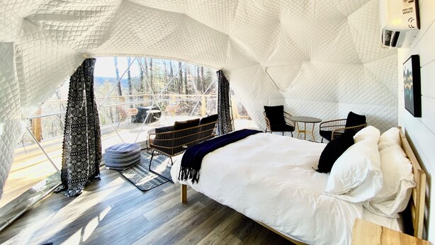 A cozy, uniquely designed room with a large bed, a small table, and a chair. The room features a distinctive geometric ceiling and large windows offering a view of the outdoors.
