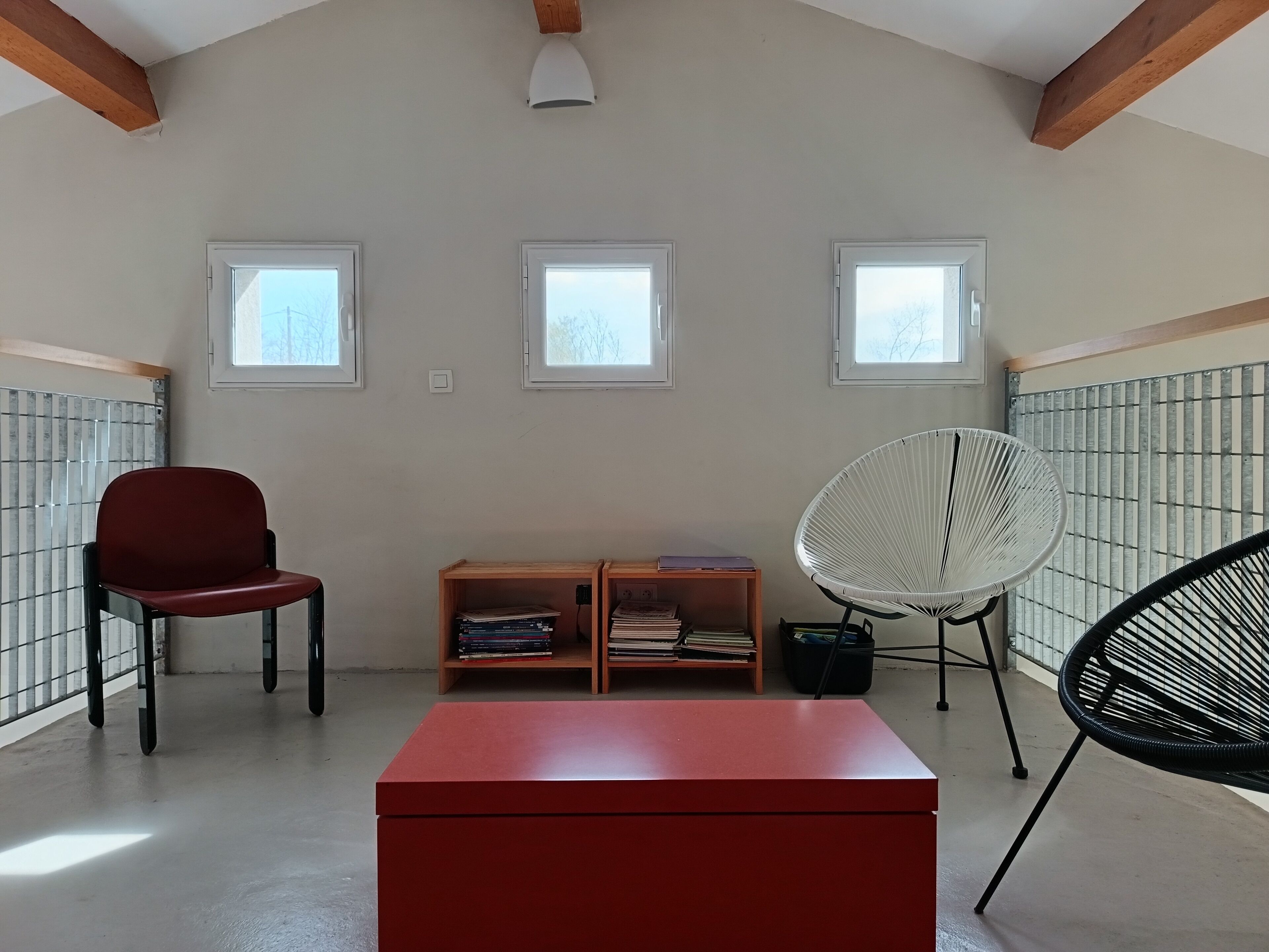 A room with a red table, two chairs, and a bookshelf. There are three windows providing natural light.
