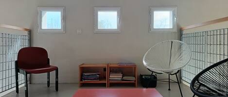 A room with a red table, two chairs, and a bookshelf. There are three windows providing natural light.