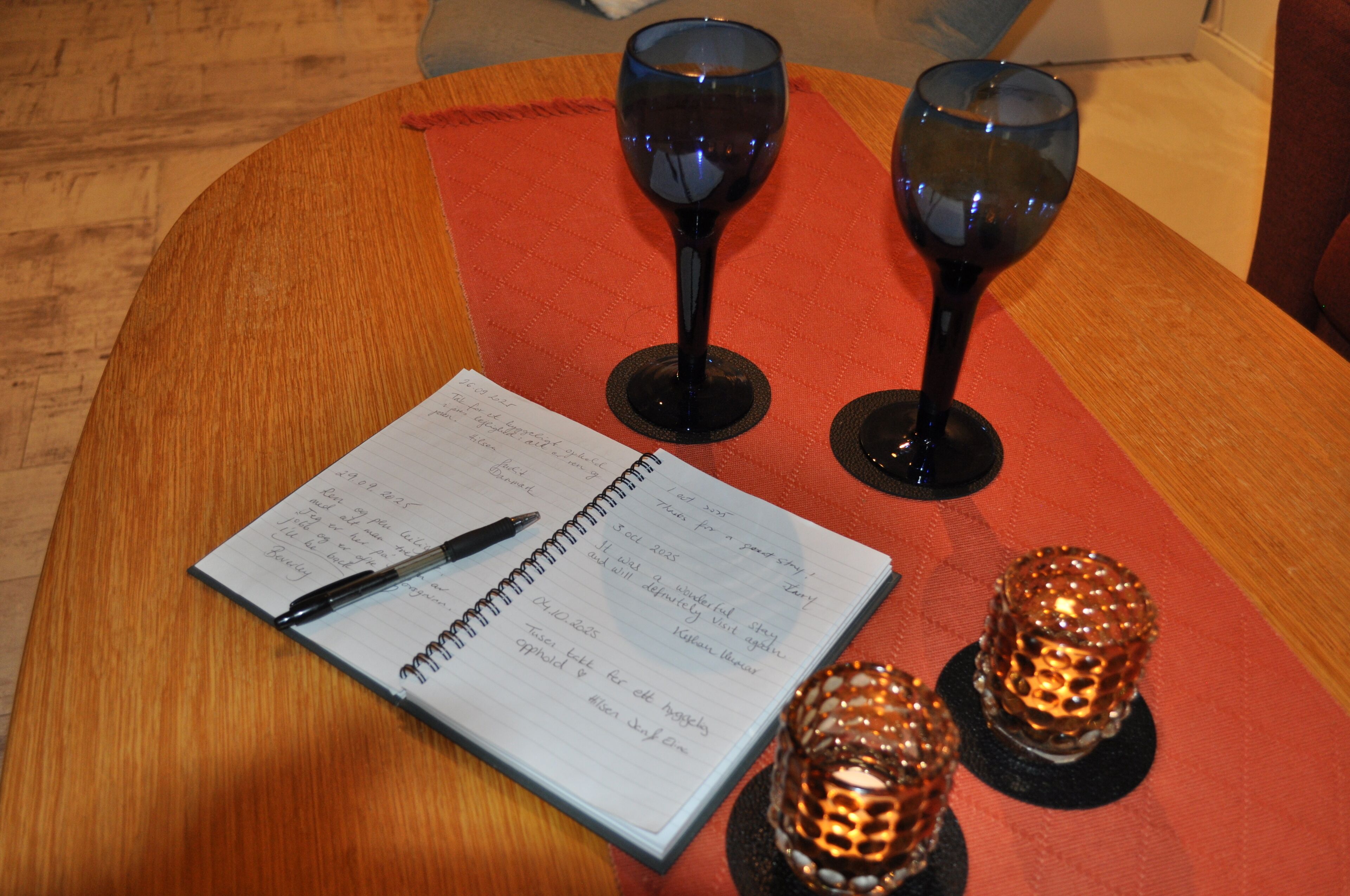 Ein Holztisch mit einem Spiralheft, zwei Weingläsern und zwei dekorativen Kerzenständern.