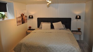 Une chambre à coucher avec un lit, deux tables de chevet, une lampe fixée au mur, une fenêtre avec une plante et un tableau encadré accroché au mur.
