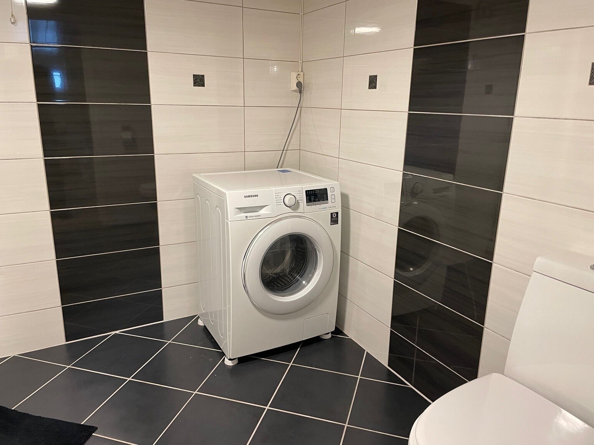 Une petite buanderie moderne, dotée d’un lave-linge à chargement frontal, de murs carrelés en noir et blanc et d’un sol carrelé de couleur noire.