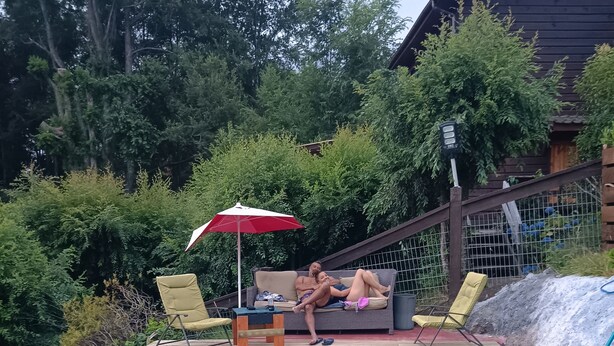 A swimming pool with a wooden deck, a red umbrella, and a couple relaxing on a sofa. - Calafquen adventure Hostel y cabañas (Conaripe)