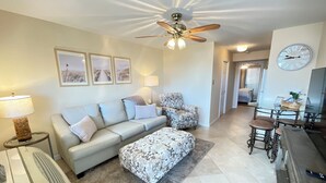 A living room with a sofa, ottoman, and a ceiling fan. There is a dining area with a table and chairs.