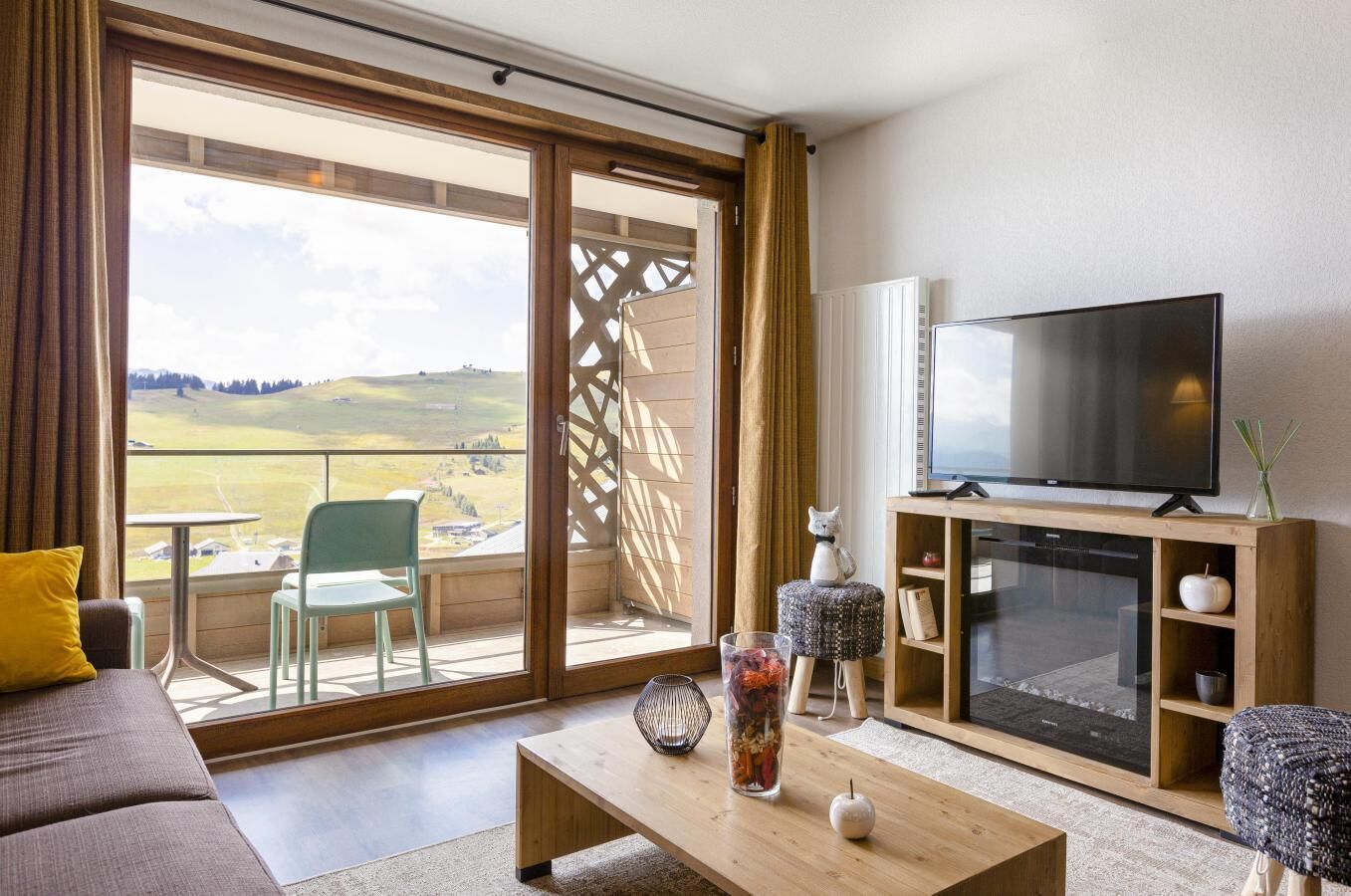 A modern living room with a large TV, a sofa, a coffee table, and a view of a green landscape through the balcony doors.