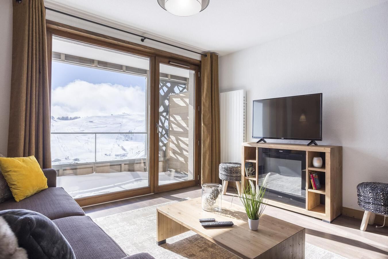 A modern living room with a large sliding glass door leading to a balcony, a flat-screen TV on a wooden stand, a sofa with a yellow pillow, and a coffee table with a plant and remote controls.
