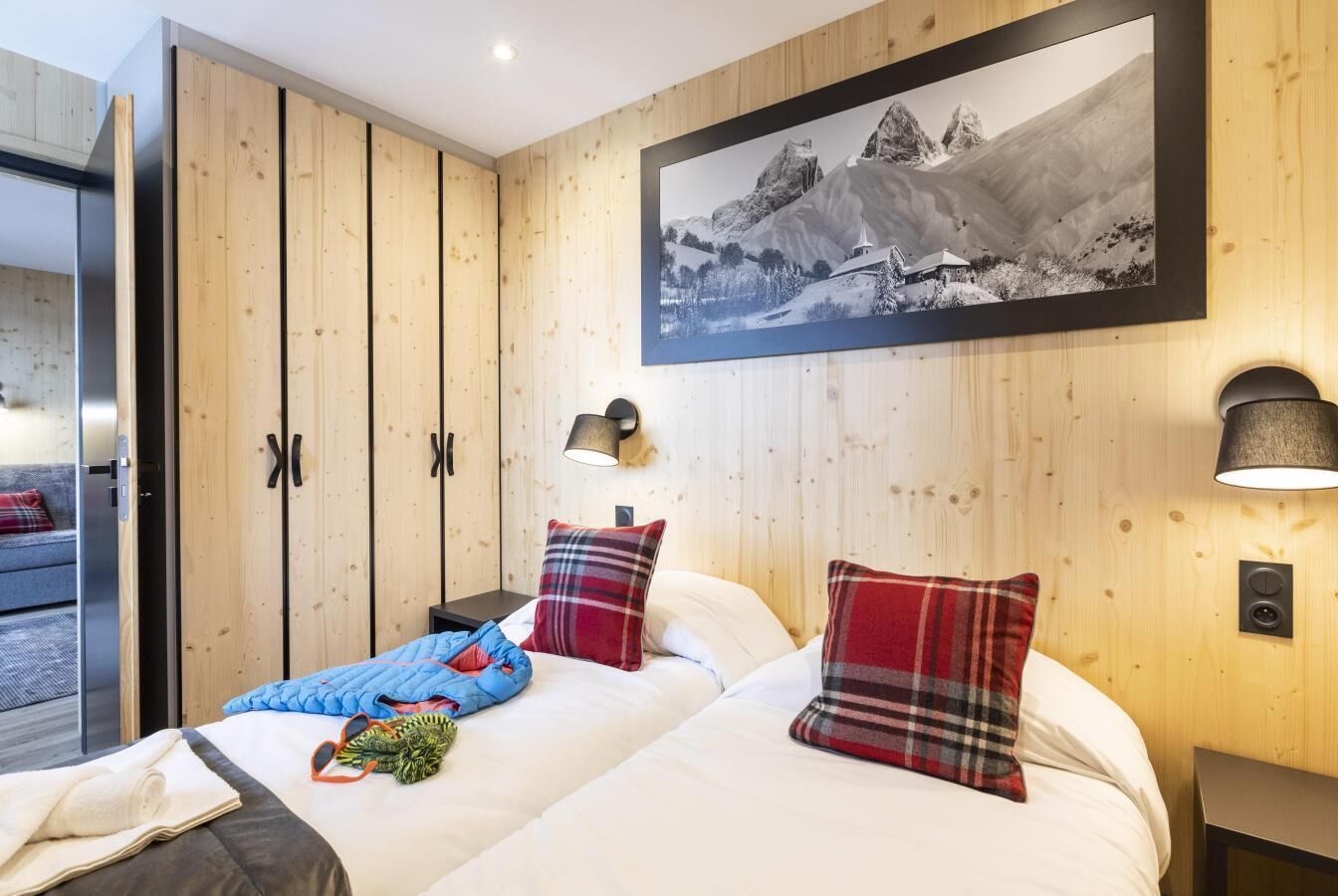 A hotel room with wooden panel walls, a bed with white bedding and plaid pillows, a mounted picture of a mountain landscape, and a nightstand with a lamp.