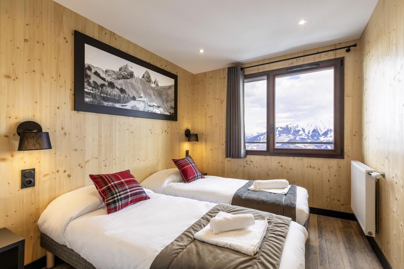 Hotellihuone, jossa on kaksi sänkyä, suuri ikkuna, josta avautuu näkymä vuorille, puupaneelit seinissä ja seinällä kehystetty taulu.