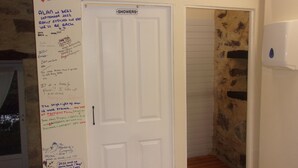 A white door with a 'SHOWERS' sign, a wall with handwritten notes, and a stone-patterned wall. - Fortified Farm 10 Berth Dormitory (La Forêt-sur-Sèvre)