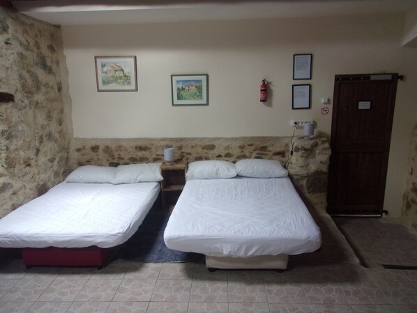 A room with two single beds, a stone wall, a wooden door, and two framed pictures on the wall. - Fortified Farm 10 Berth Dormitory (La Forêt-sur-Sèvre)