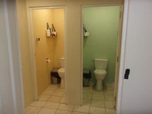 A narrow hallway leading to two bathrooms, one with a toilet and a trash bin, and the other with a toilet and a small trash bin. - Fortified Farm 10 Berth Dormitory (La Forêt-sur-Sèvre)