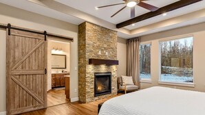 A bedroom with a large bed, a stone fireplace, a sliding barn door, and a view of a snowy landscape. - The Dreamland House - Central, Stylish, Spacious (Wasilla)