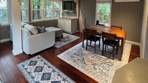 Een ruime woonkamer met houten plafond, een bank, een eettafel met stoelen en een aan de muur bevestigde televisie.