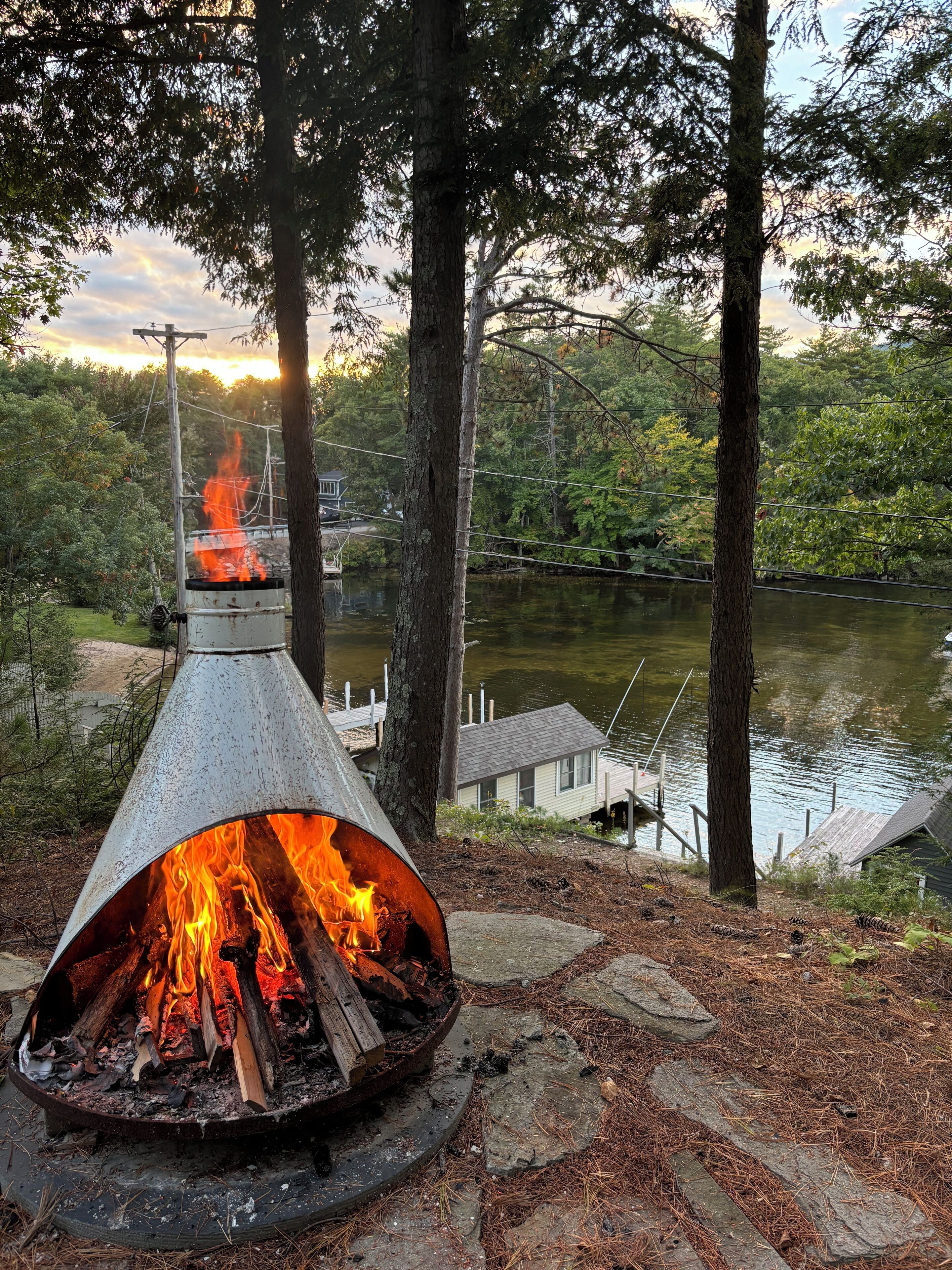 Ein Lagerfeuer in einer Metallfeuerstelle, umgeben von Bäumen und Blick auf einen See.
