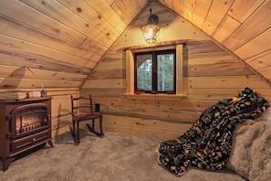 A cozy cabin interior with a wooden ceiling, a fireplace, a rocking chair, and a window with a view of trees.