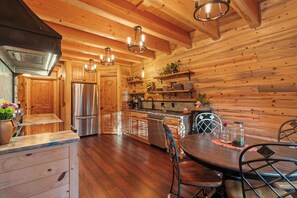A wooden interior with a kitchen area featuring stainless steel appliances, a dining table with chairs, and a ceiling with exposed beams and pendant lighting.