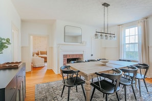 Un comedor con mesa de madera y sillas negras, una chimenea y un ventanal con cortinas.