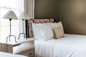 Una cama bien hecha con sábanas blancas, una mesita de noche de madera con dos lámparas y un cabecero estampado con motivos florales.